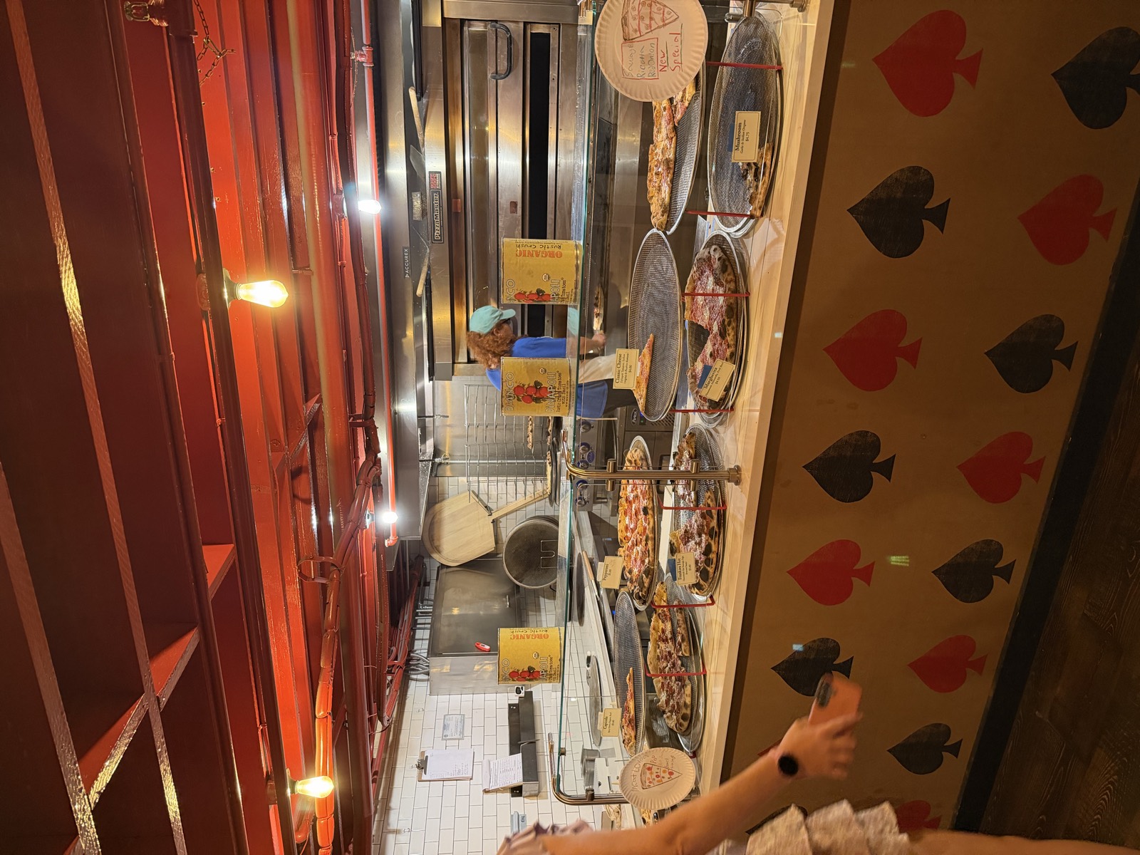 The slice counter at Frankies Pizzeria, with whole pies on trays in front of the oven and a red and black spade mural