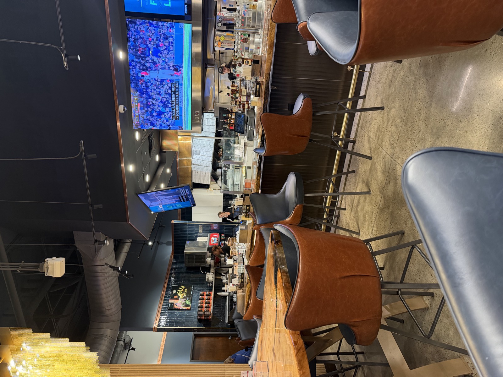 The bar at NY Pie, with leather stools, TVs over the counter and the pizza line behind it