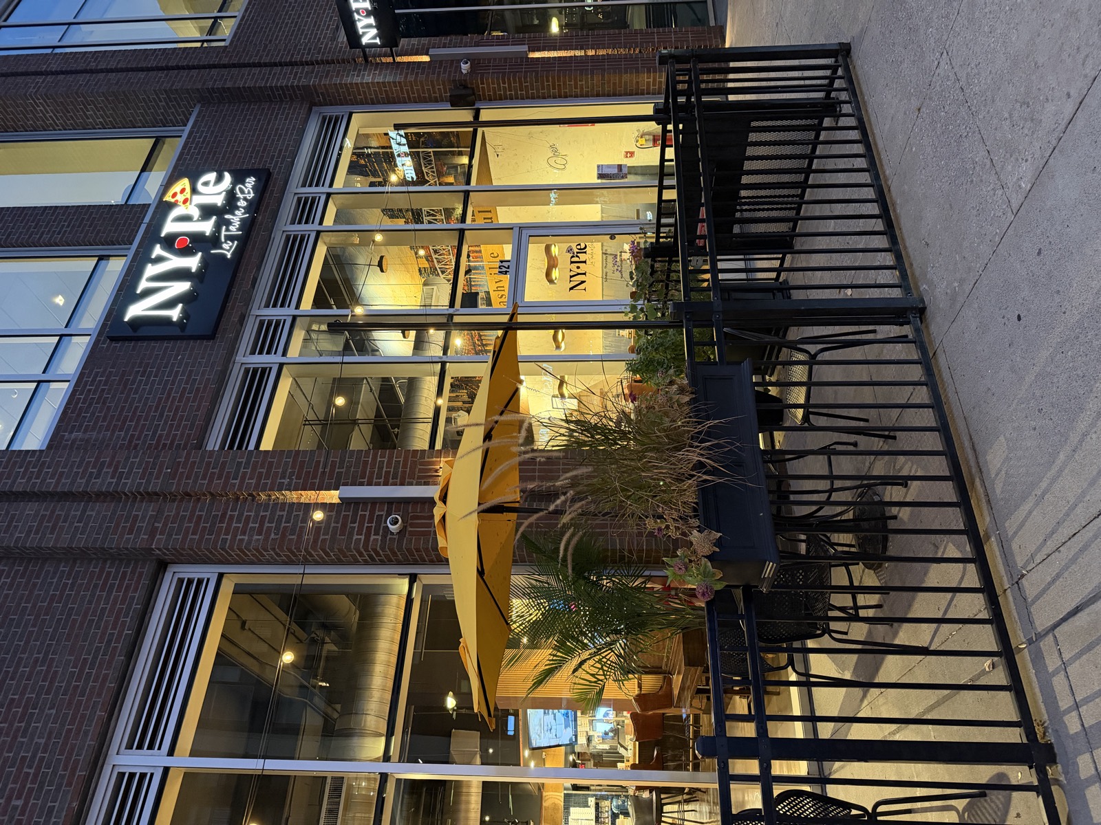 NY Pie's entrance at Capitol View in the evening, with a yellow patio umbrella and fenced patio seating out front