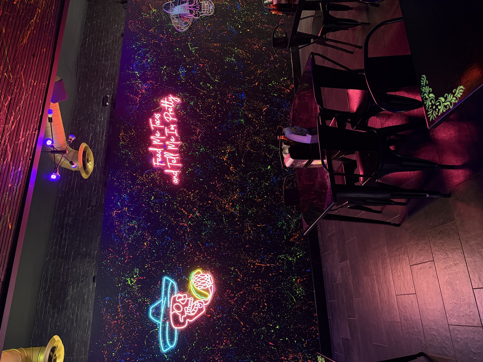 The splatter-painted wall at Talkin' Tacos, lit by neon signs including Feed Me Tacos and Tell Me I'm Pretty